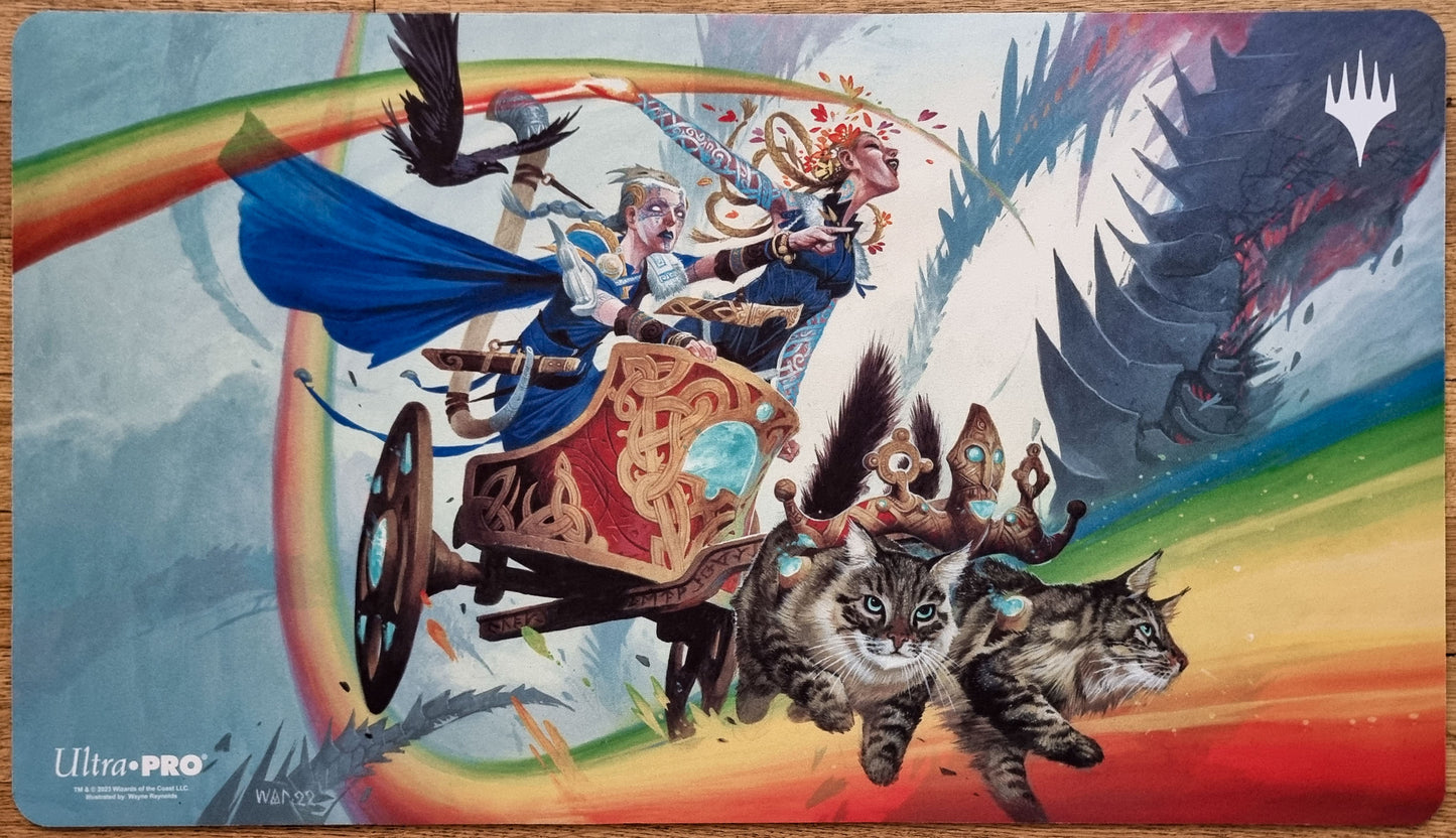 Inga and Esika MTG playmat by Wayne Reynolds showing two figures in a rainbow chariot pulled by cats, with ravens and a dark mechanical threat behind them.