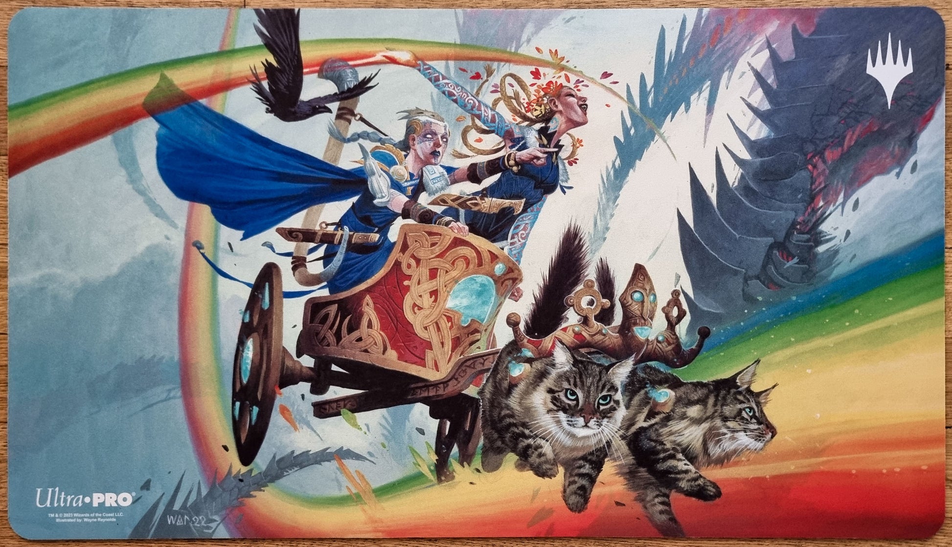 Inga and Esika MTG playmat by Wayne Reynolds showing two figures in a rainbow chariot pulled by cats, with ravens and a dark mechanical threat behind them.
