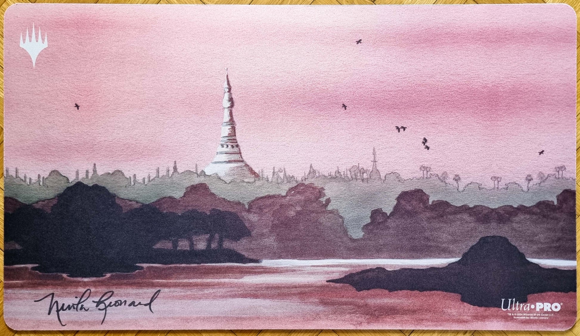 Signed Karakas MTG playmat by Nicola Leonard with the newer Magic logo, showing a pink landscape, distant tower, water, and silhouetted scenery.