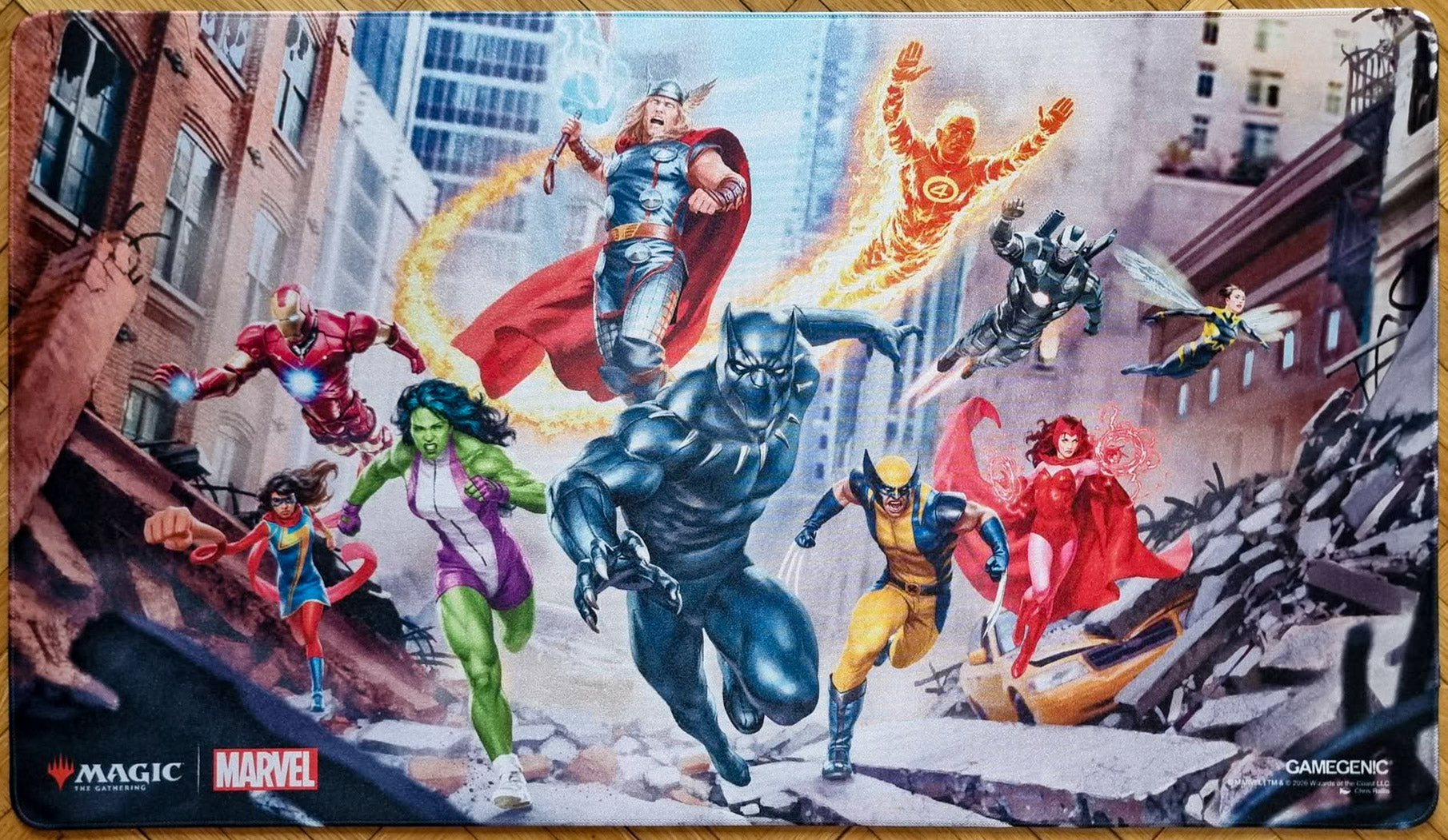 Magic: The Gathering x Marvel shiny playmat featuring Chris Rallis’s Marvel Super Heroes key art, with Black Panther, Thor, Iron Man, Wolverine and other heroes charging through city ruins.