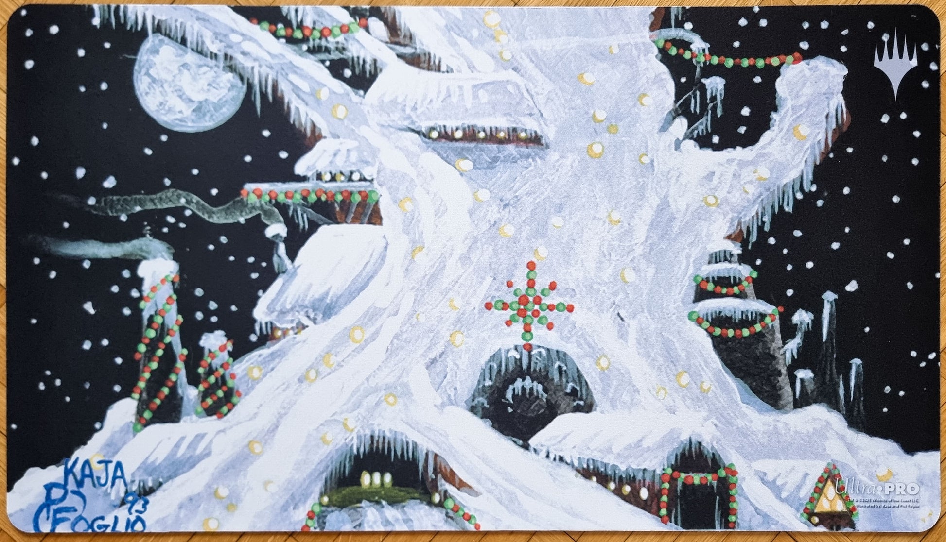 Winter Mishra’s Factory MTG playmat by Kaja and Phil Foglio, showing the snow-covered Antiquities factory decorated with festive lights under falling snow.
