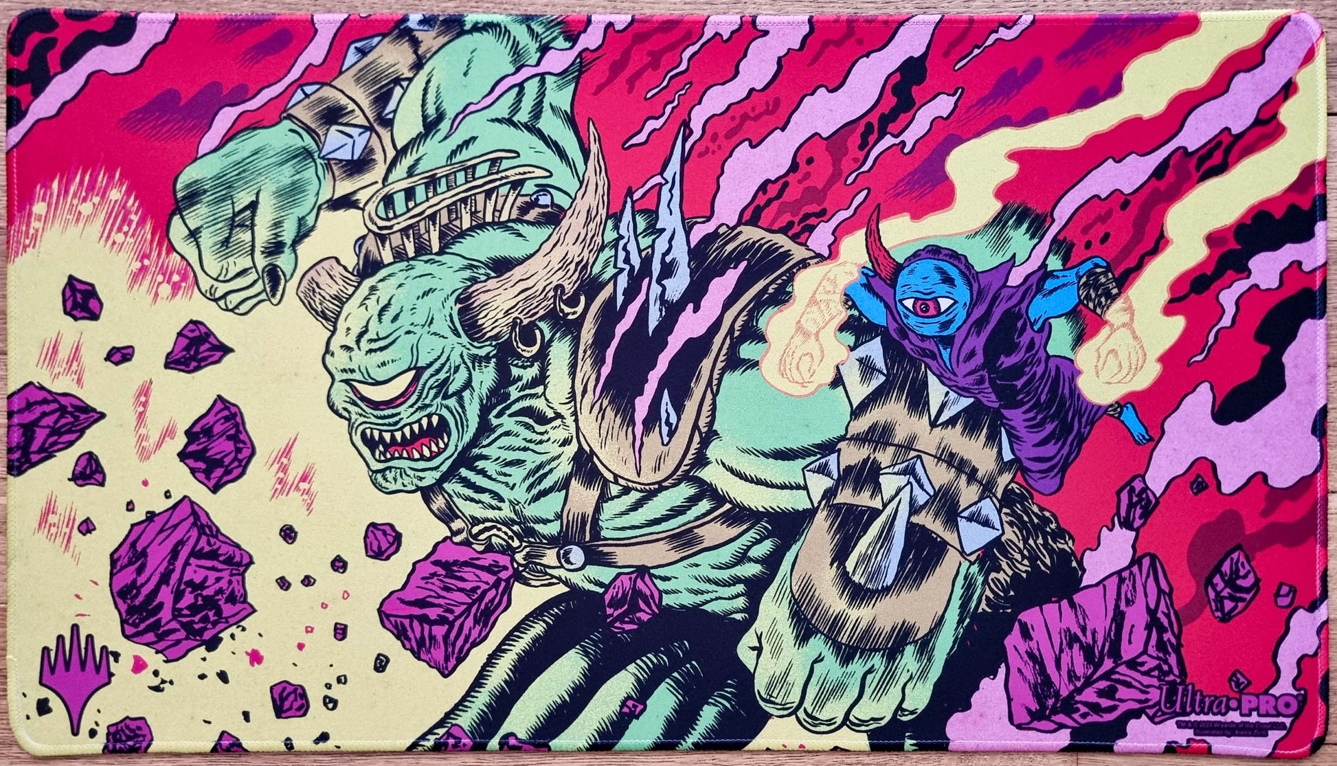 Stitched Okaun and Zndrsplt MTG playmat by Alexis Ziritt showing a green cyclops and blue homunculus in a vivid comic-style chaos scene.
