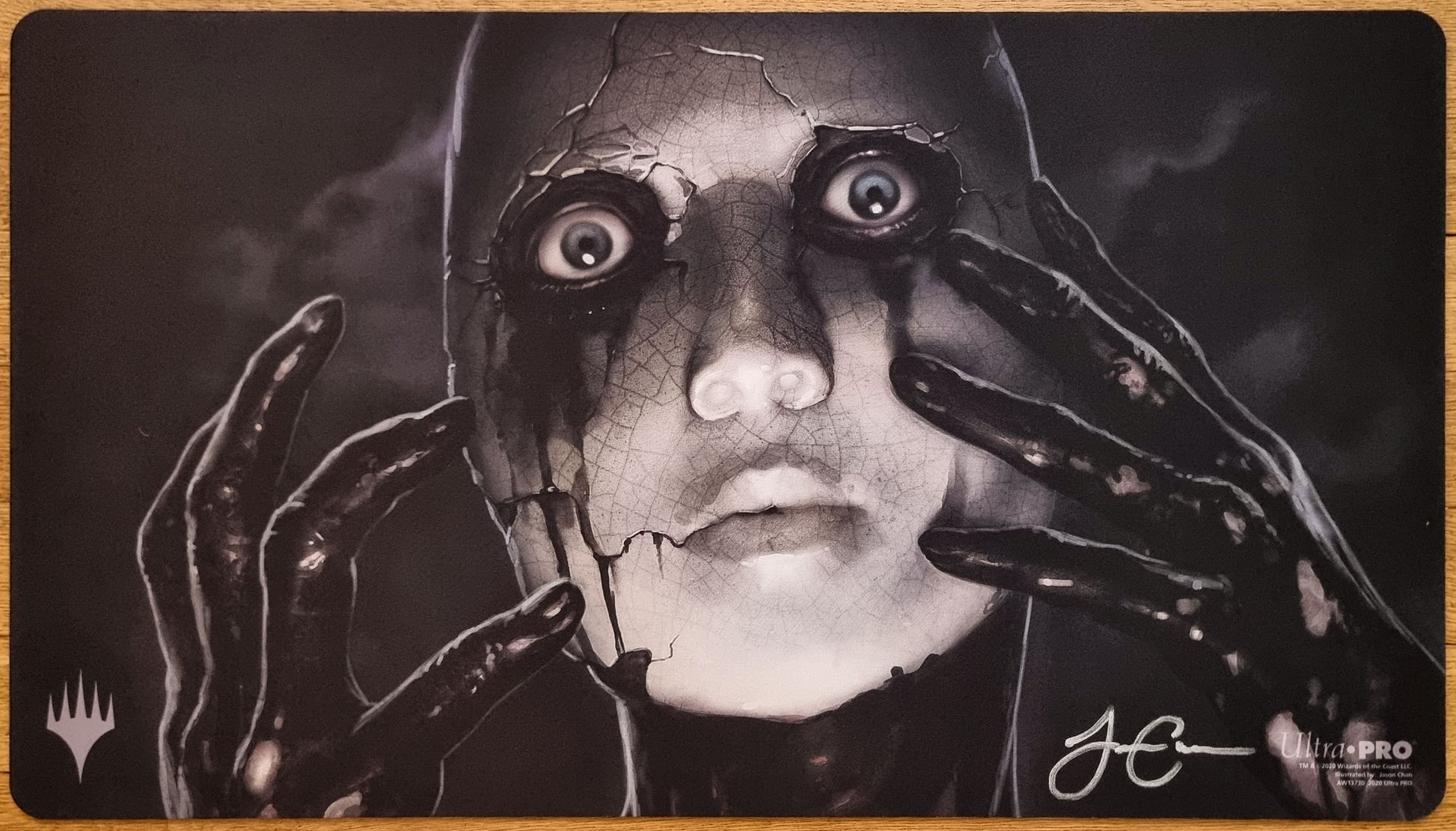 Signed Phyrexian Unlife MTG playmat by Jason Chan showing a cracked porcelain-like face with dark hands in a monochrome horror scene.