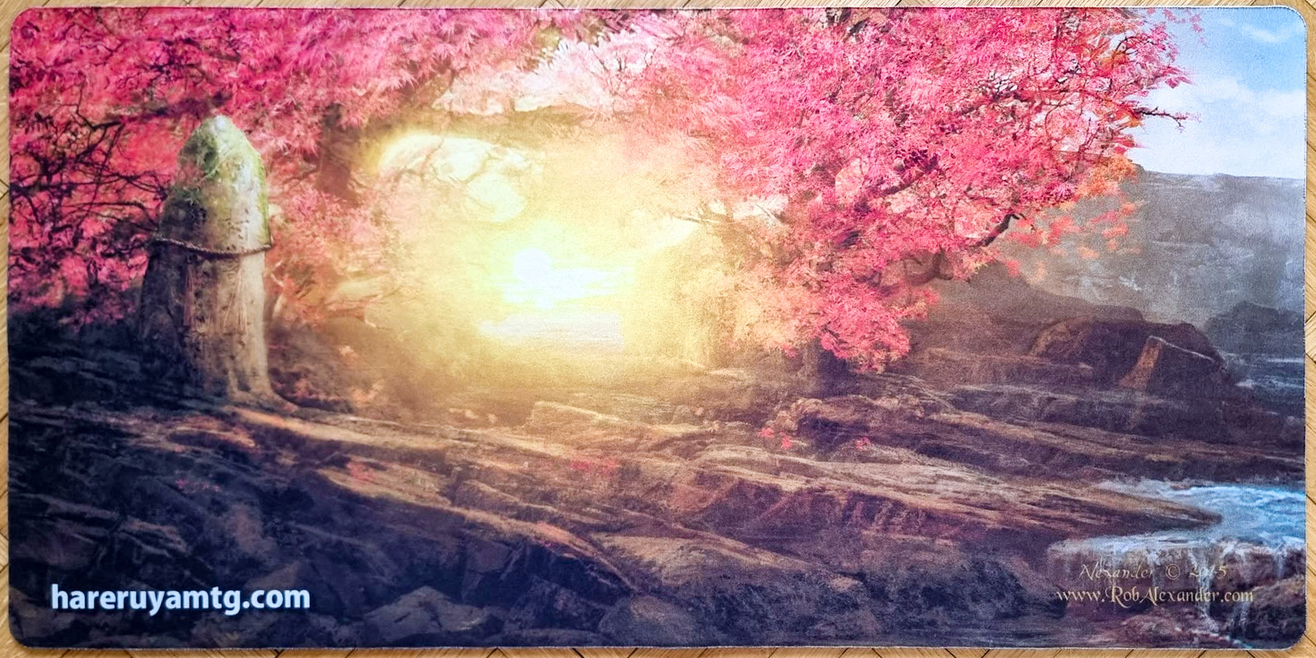 Rob Alexander Spring Sakura playmat from Hareruya’s 2015 Four Seasons in Japan series, showing pink cherry blossoms, a shimenawa-wrapped stone, and glowing morning light.