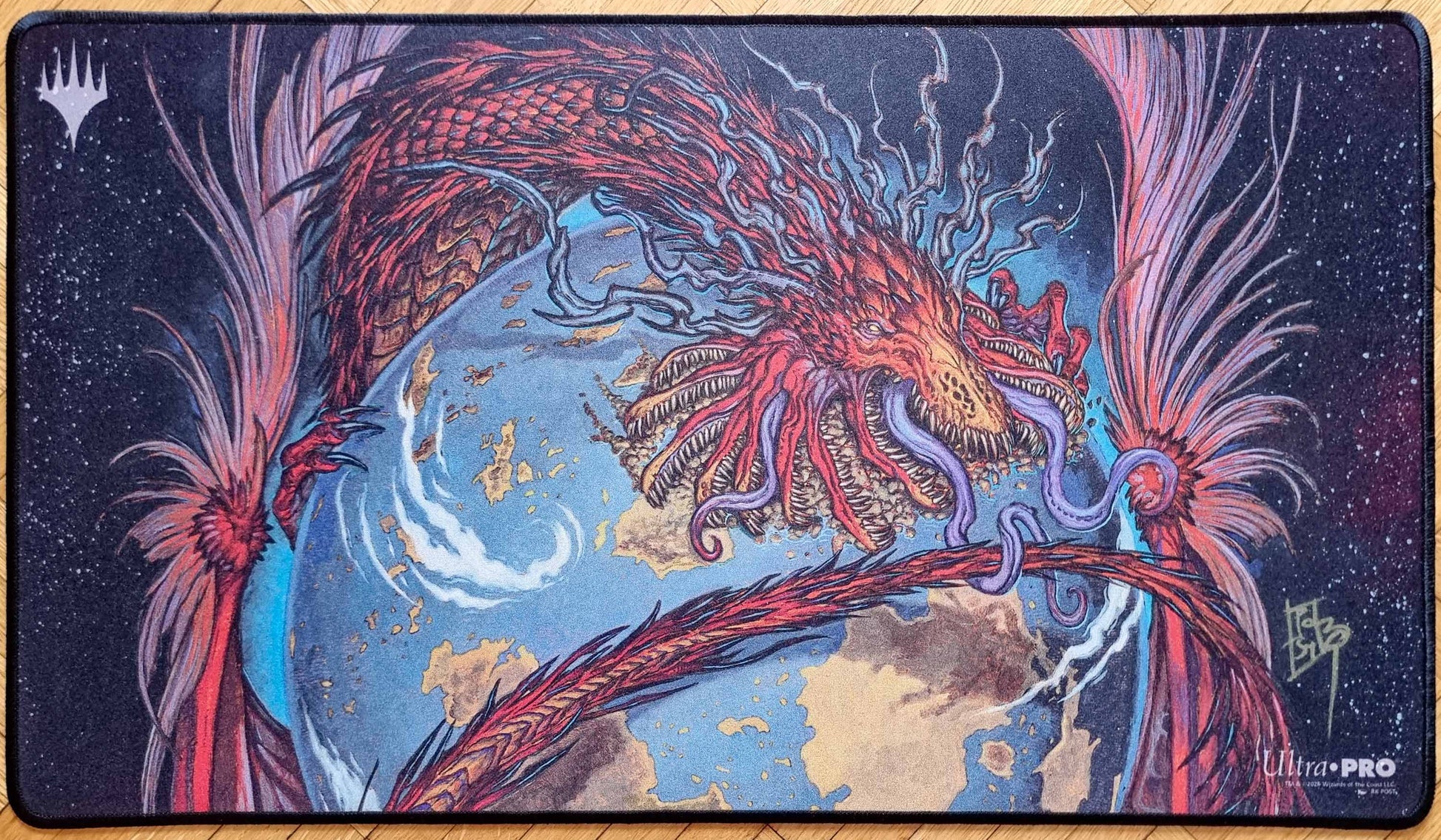 Signed Worldgorger Dragon MTG playmat featuring rk post’s Dominaria Remastered artwork, showing a red dragon coiled around a blue world with stitched black edges.